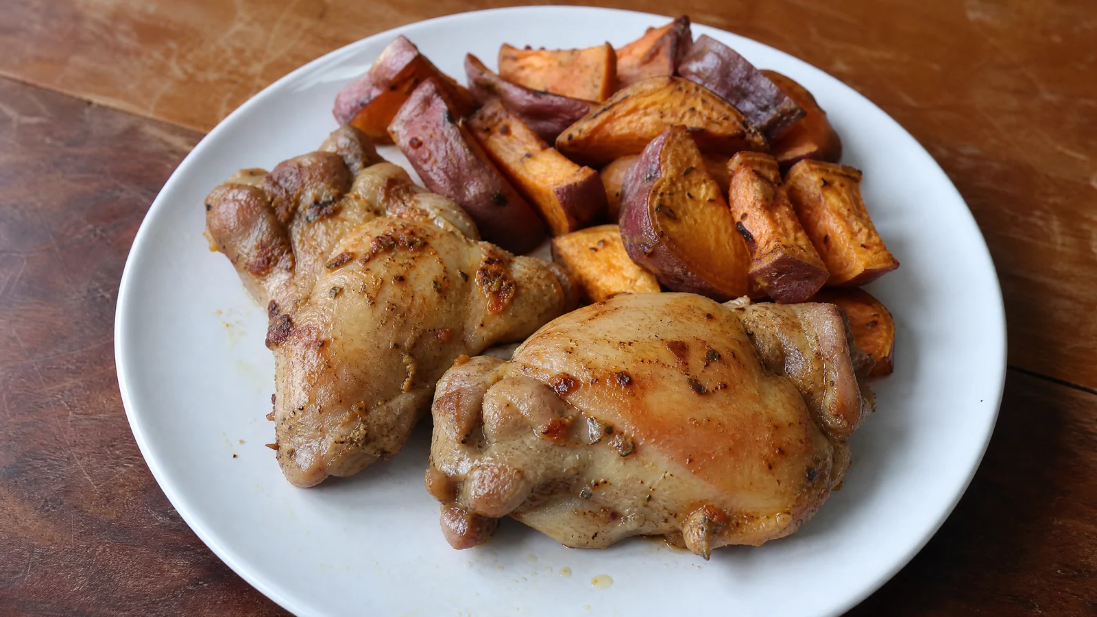 Prato branco com duas sobrecoxas de frango sem pele douradas e temperadas, ao lado de pedaços de batata-doce assada com casca roxa, sobre mesa de madeira.