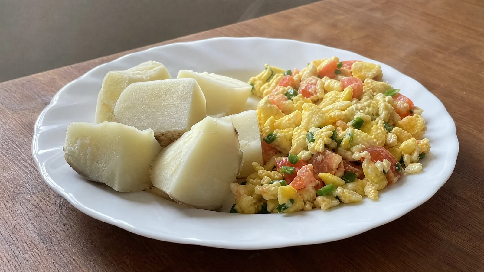 Prato branco com pedaços de inhame cozido de polpa clara e ovo mexido com tomate picado e cebolinha, sobre mesa de madeira.